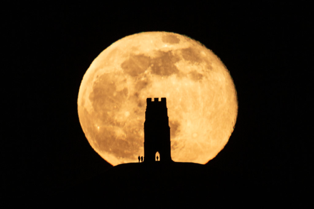 silhouette of Glastonbury Tor and three small figures with the Full Moon in the background appearing almost twice the size 