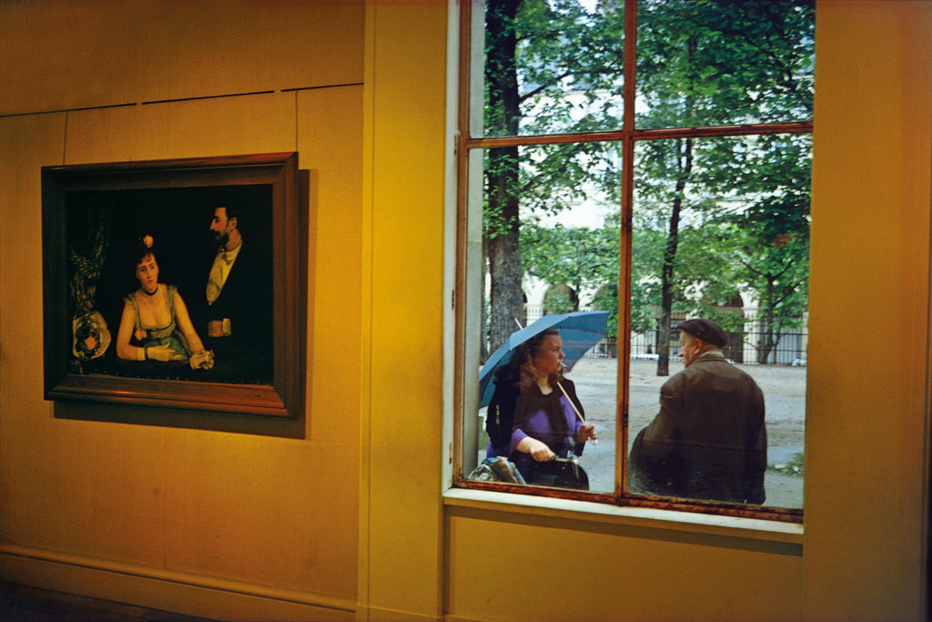 Joel Mayerowitz, a gallery interior with yellow walls and a painting of a couple onn the wall, through the window of the gallery we see a real life couple