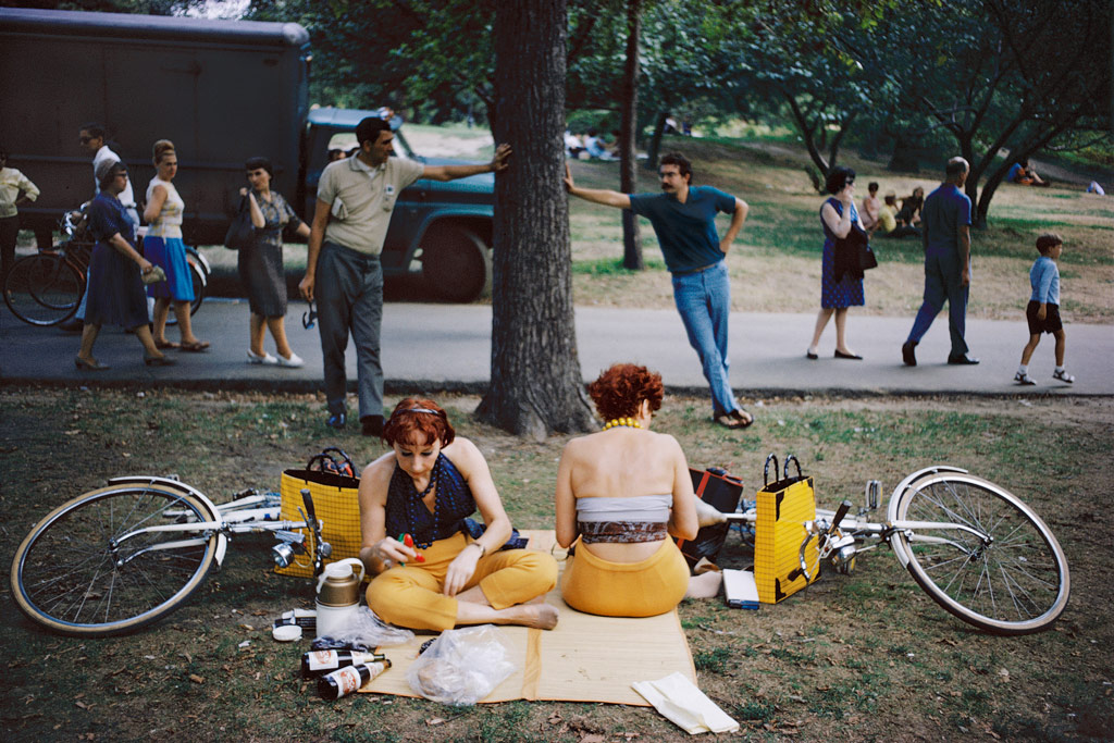 Joel Mayerowitz, two women having a picnic in a park, their mustard yellow trousers and bags create a striking contrast with their surroundings, and the two men lenaing against a tree next to them