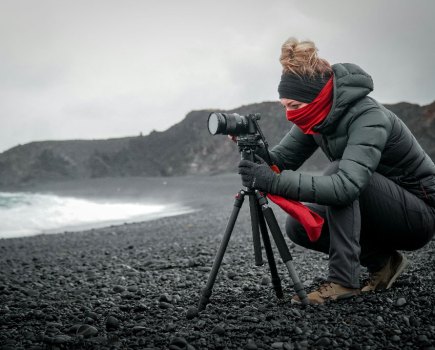 a woman taking a photo with a camera on a tripod, on a rocky grey beach wearing grey clothes and a red scarf