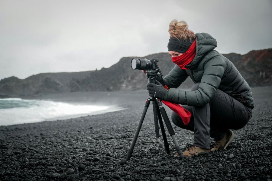 a woman taking a photo with a camera on a tripod, on a rocky grey beach wearing grey clothes and a red scarf
