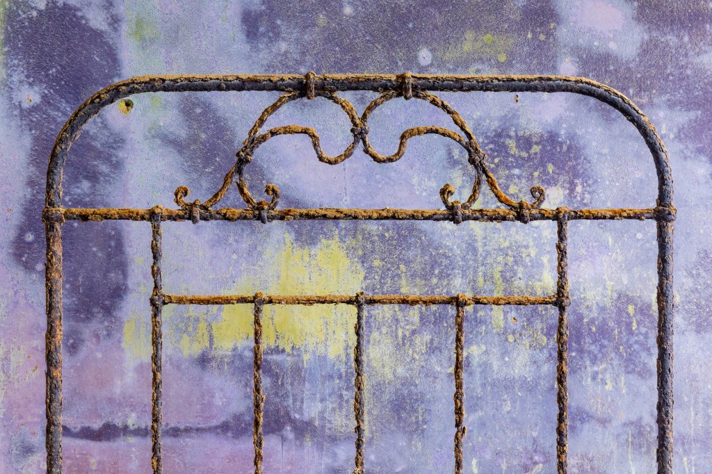 A rusty bed frame against purple and yellow chipped wall paint