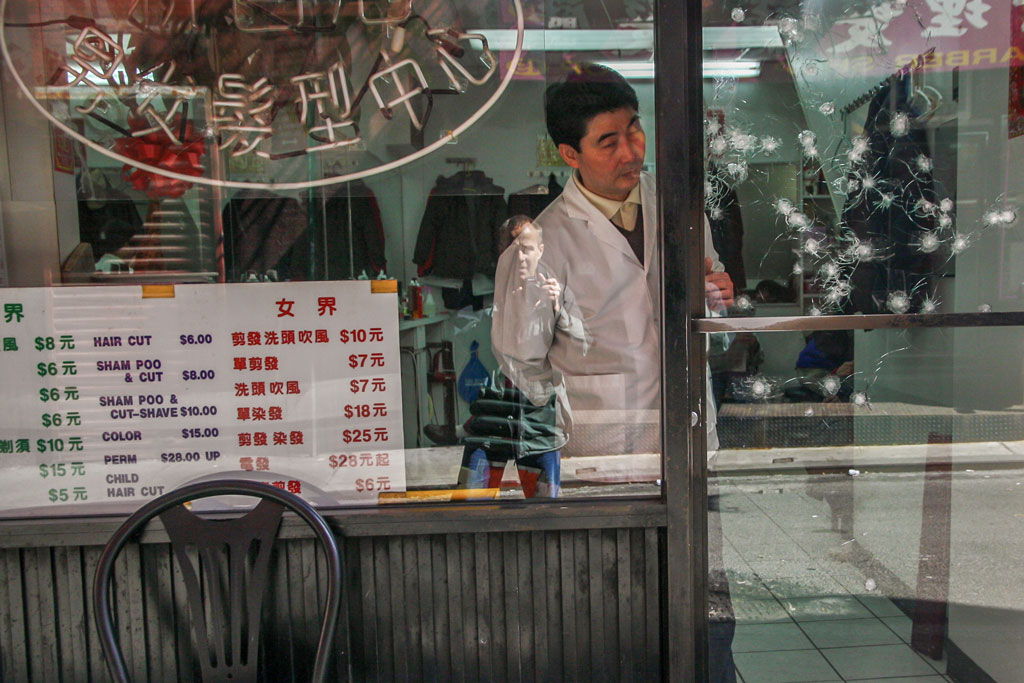 New. York, dry cleaners worker closes the front door which glass door which seems yo have been shot at multiple times. the photographer reflected in the window