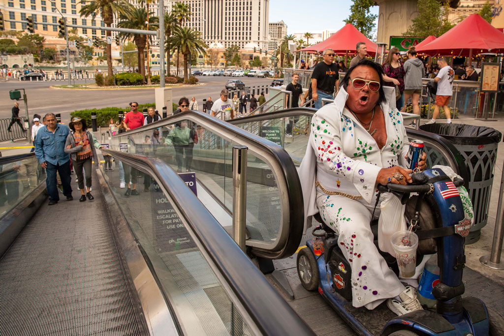 A man dressed as Elvis rides a mobility scooter up a ramp while shouting at the camera, street photography America