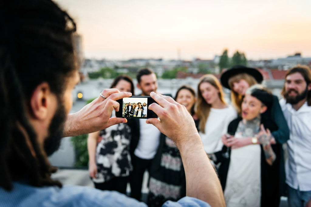 Young man taking pictures of friends with his smart phone on the roof top