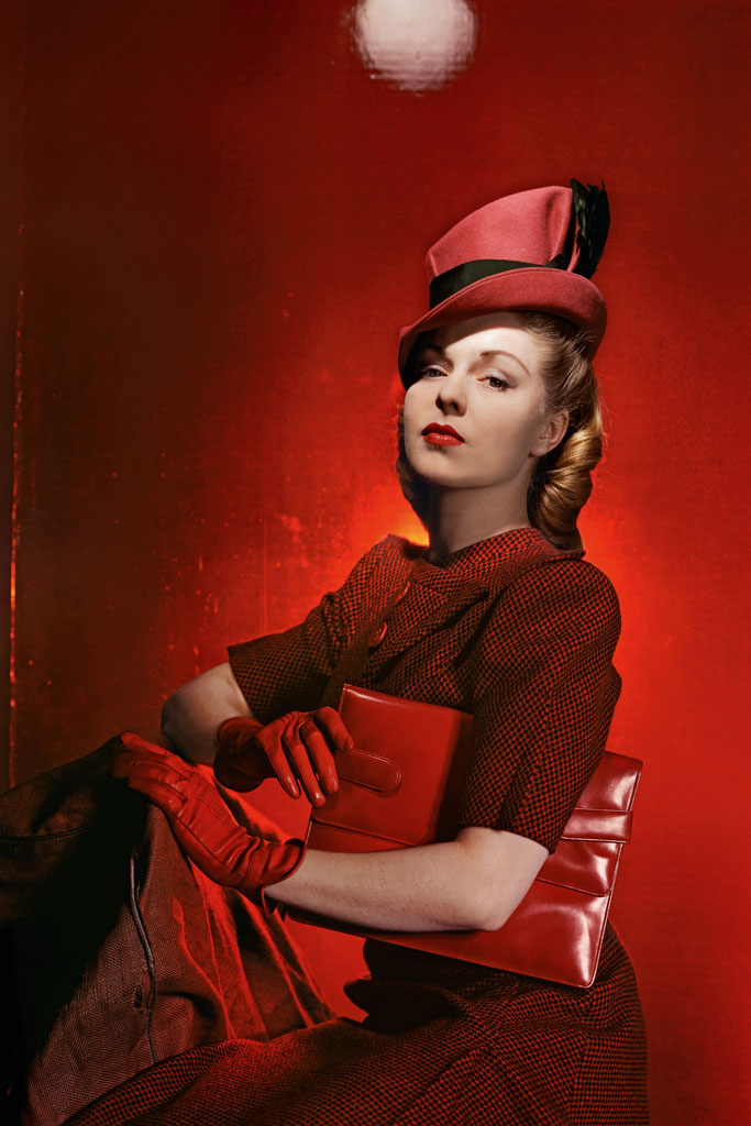 A woman in red haute couture dress with gloves  and a hat poses in front of a red backgroud