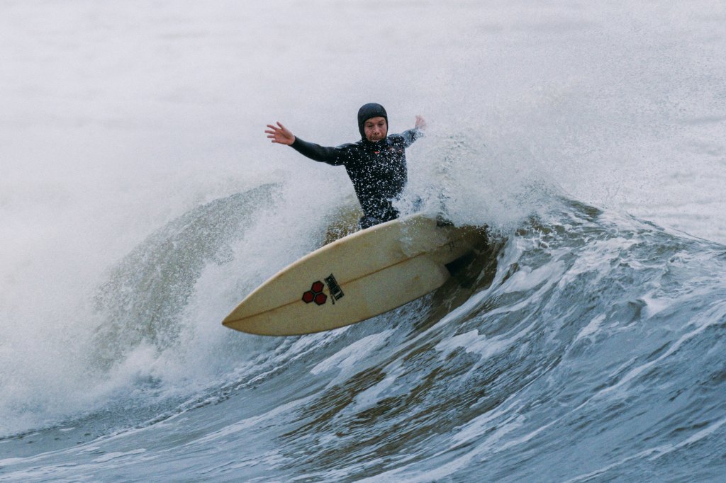 As surfer in black wetsuit on a white board cuts through a wave