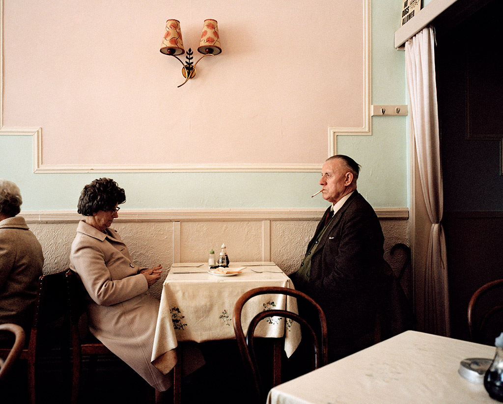 man and woman at a table in a restaurant sat infront of pastel pink wall i am martin parr film