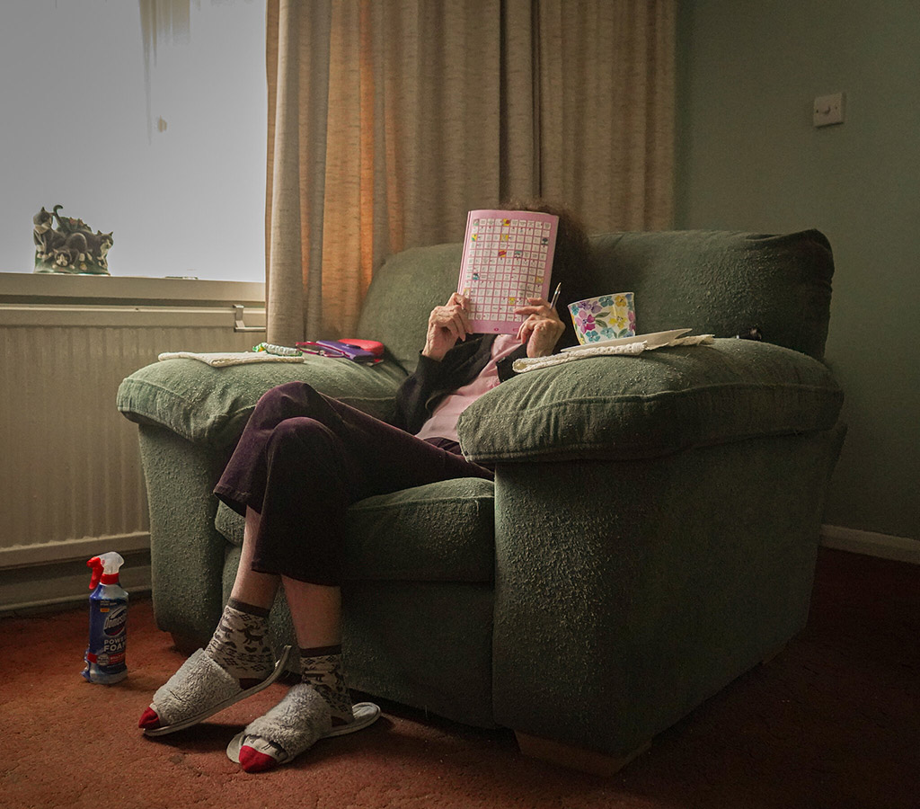 older woman in a green armchair with puzzle book over face