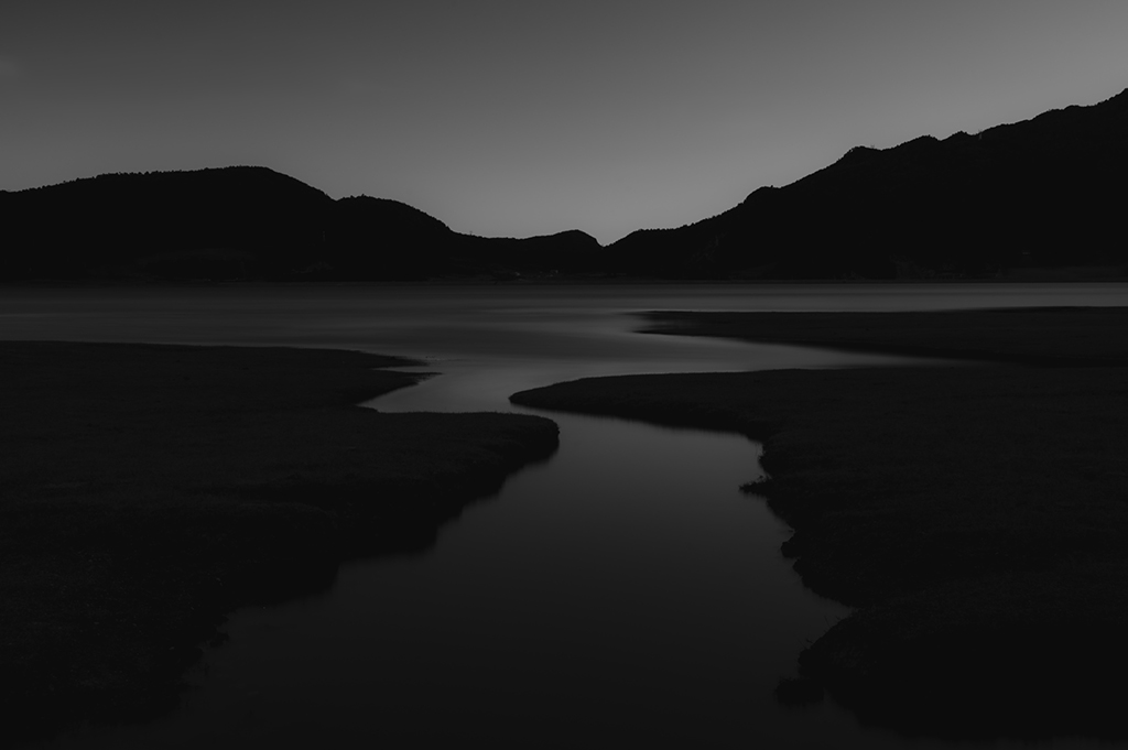 black-and-white shot of a stream meandering through grasslands into a lake