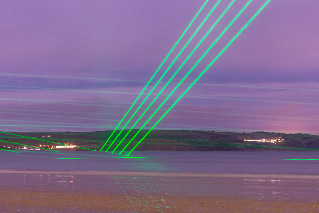 Five rays of green laser lights brake over the surface of the water in a pink coloured sunset over the sea