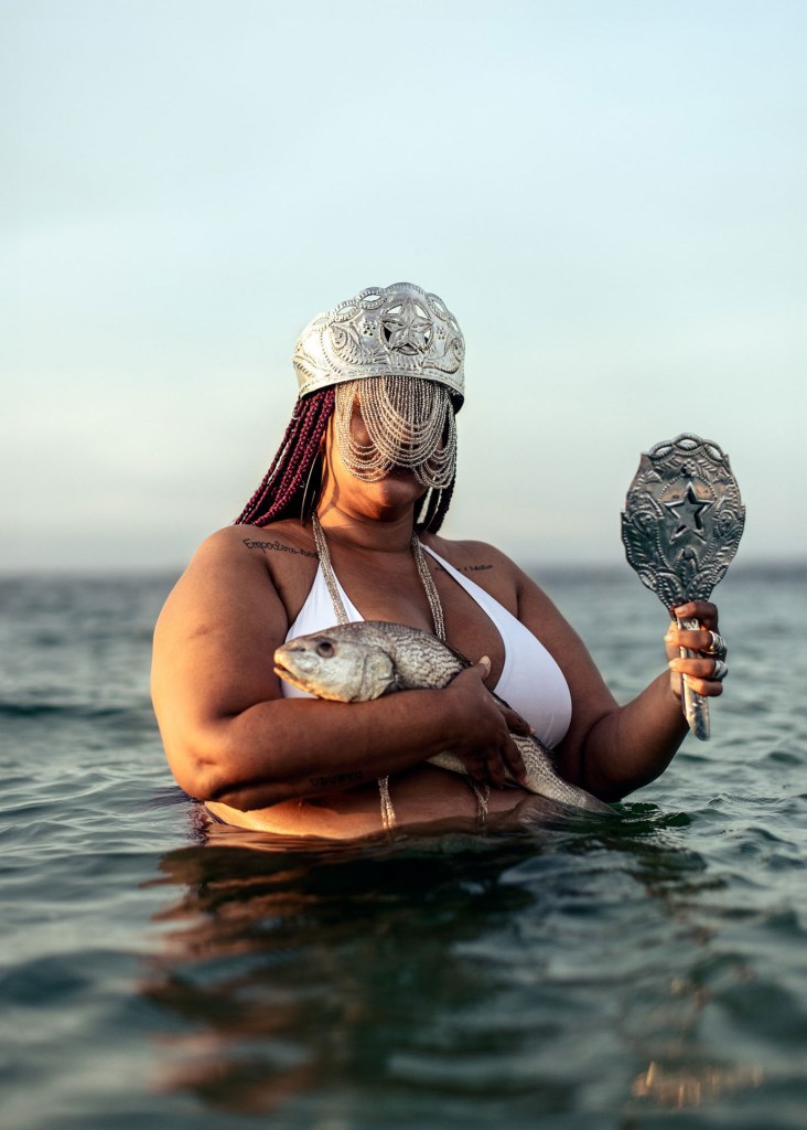 The portrait shows Candomblé practitioner Samara Souza making an offering to Yemanjá, the Yoruba deity of the sea waters. Yemanjá's name means "the mother whose sons are fishes" and she is the most popular orixá in Brazil. Due to syncretism, in Brazil she is represented by an image of a white, skinny woman, while in Africa her image shows a corpulent woman with big milking breasts.