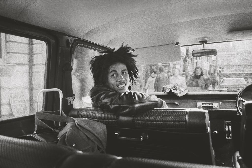 Black and white photograph of Bob Marley in the passenger seat of a van with his body partially turned towards the back of the vehicle.