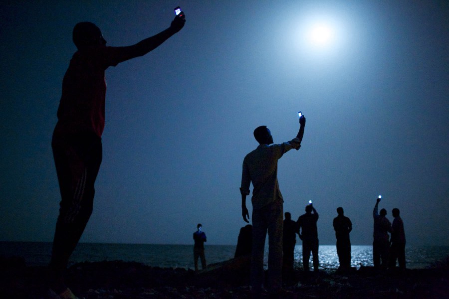 African migrants on the shore of Djibouti City raise phones in an attempt to catch an inexpensive signal from neighboring Somalia. Image Credit: John Stanmeyer, USA, VII SINGLE USE ONLY