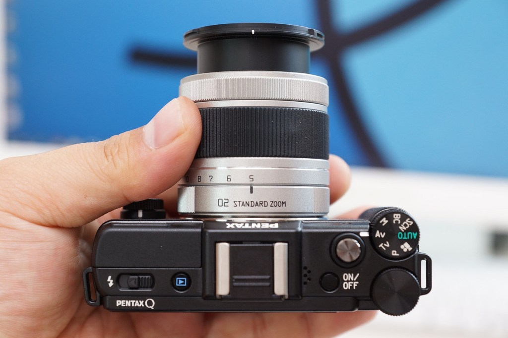 A photograph of hands holding the Pentax Q with an attached telephoto lens, indicating how small they are