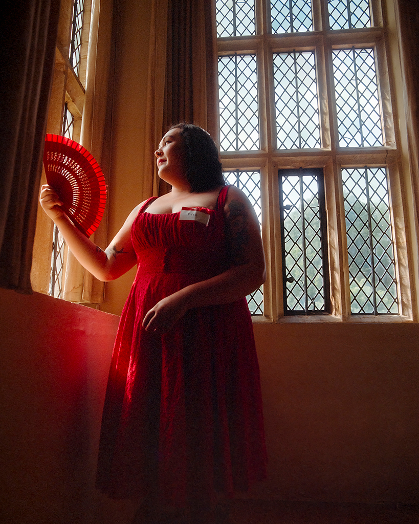 young woman posing by a window in a long red dress with red fan