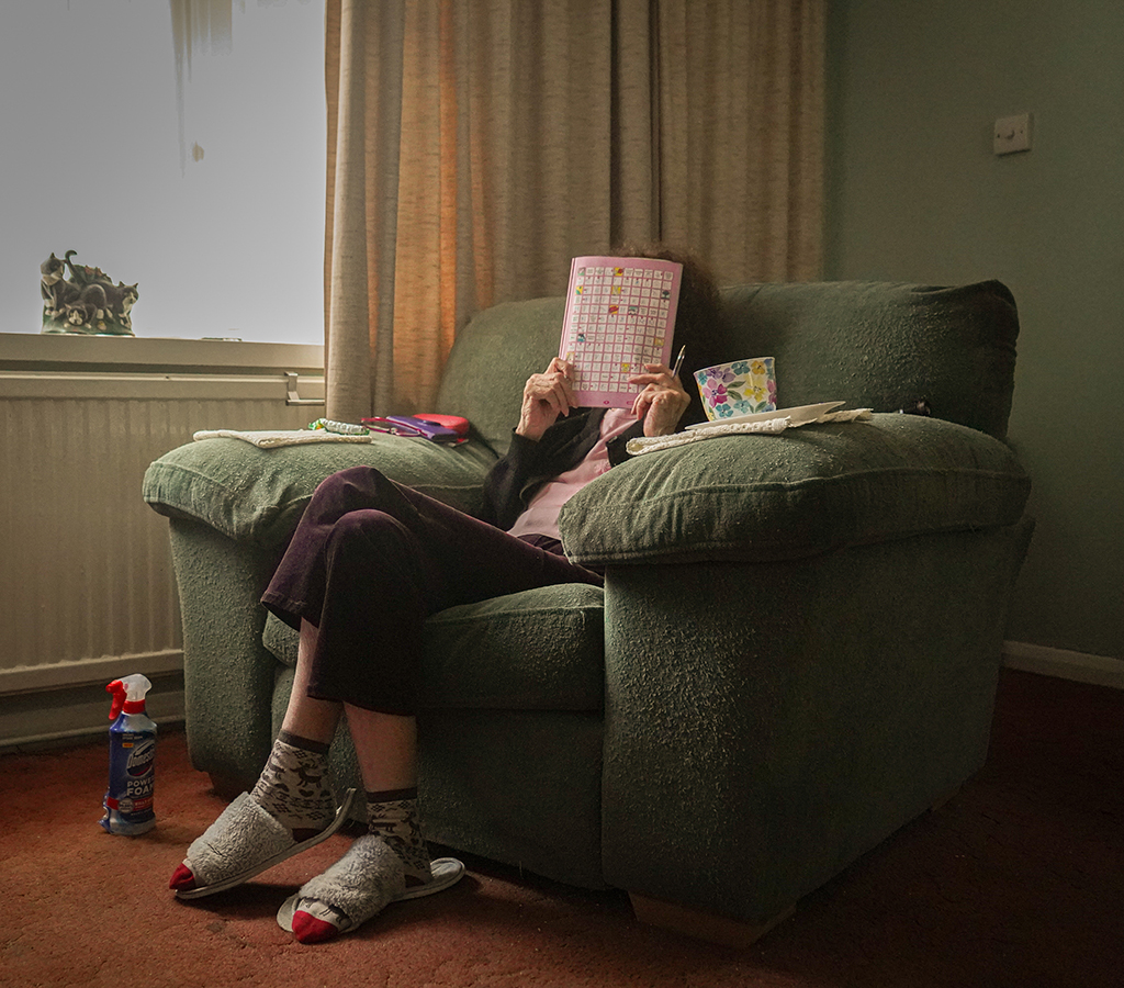 photo of a woman sat on the sofa with crossword book held up to her face