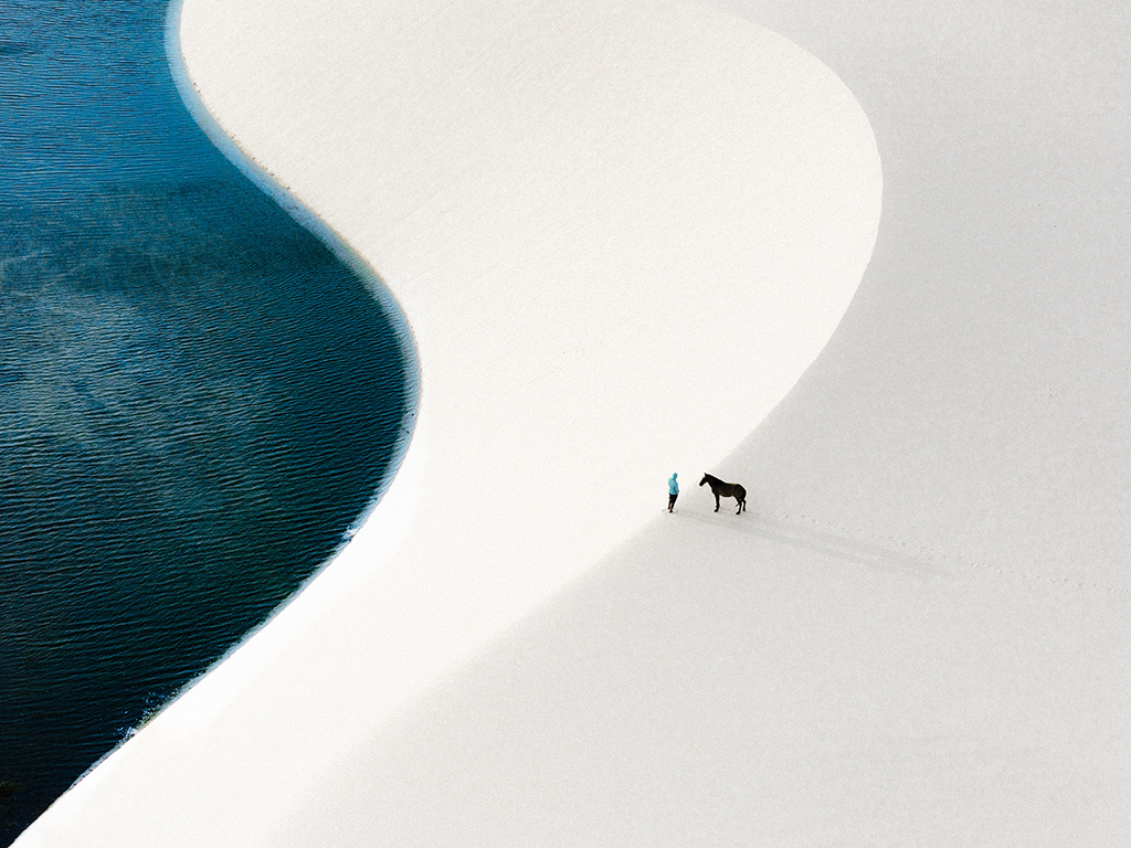 aerial photo of person stood with horse on sand dune near water