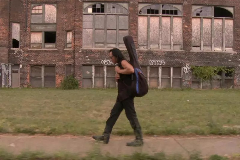 Screenshot from 2012 docuemntary Searching for Sugar Man, showing vintage footage of a man walking holding a guitar.