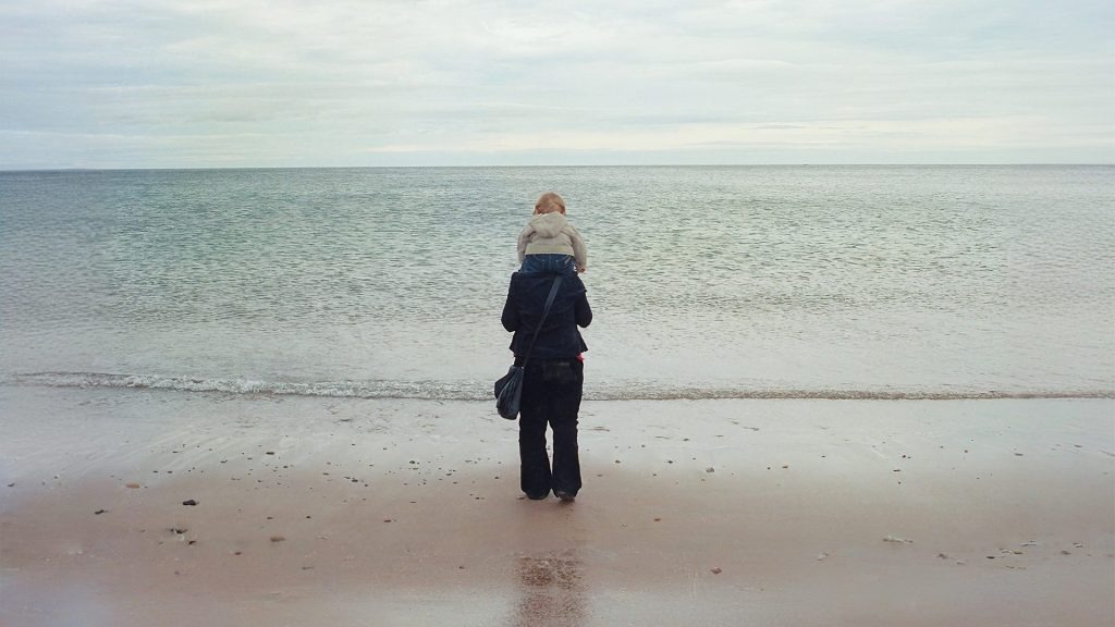 Still image from the film 'Motherboard', shot on iPhone, showing a mother with her son on her shoulders, standing on the beach looking into the sea