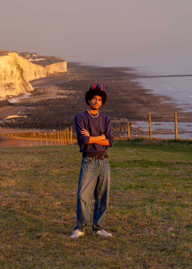 portrait of a man with multicoloured afro combs in his hair stood on a coastline