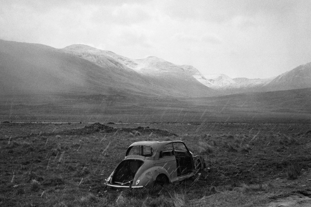 Black and white photograph of an old car in a desolate field with snow-covered mountains in the background.