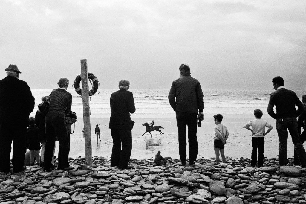 Black and white photograph showing a group of people standing on a rocky shore facing the ocean. In the distance, a person can be seen riding a horse along the water.