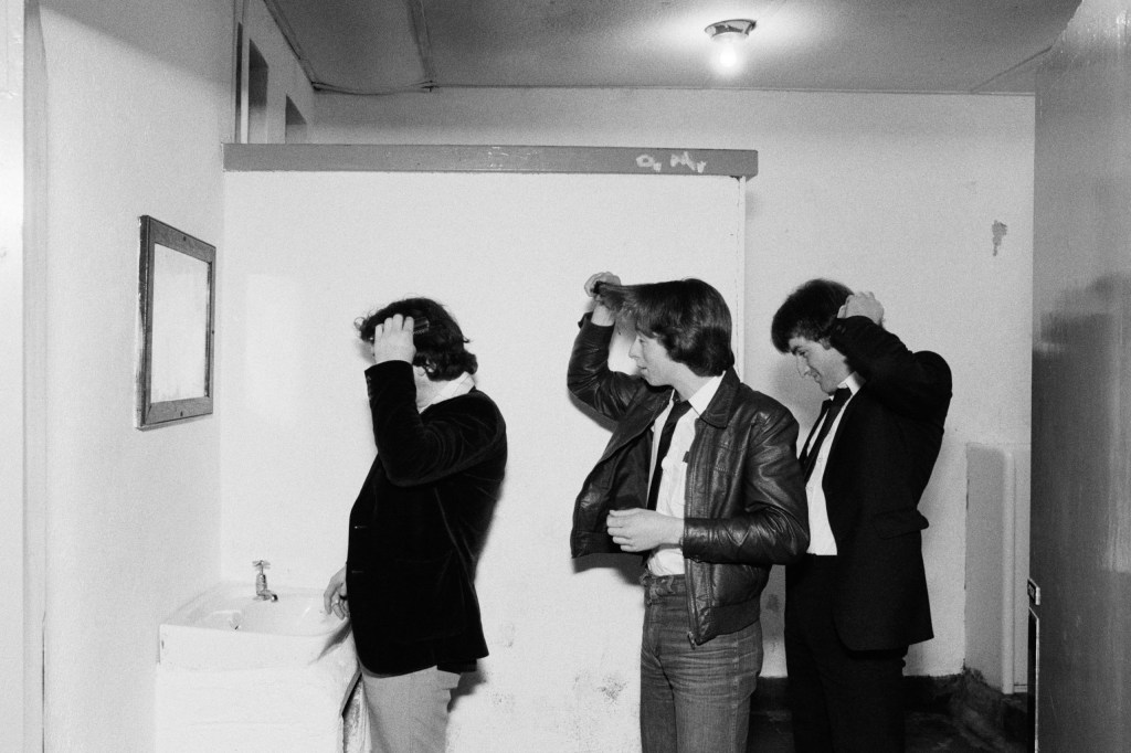 Black and white photograph of three men standing in front of a small mirror above a sink as they each attempt to fix their hair.