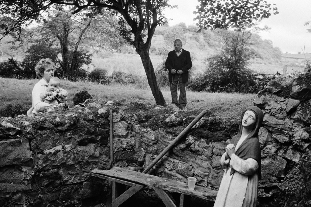 Black and white photograph depicting an outdoor setting. Behind a low stone wall, a woman sits hold- ing a baby and the man stands next a tree. A statue of Mary stands near a wooden bench in front of the wall.