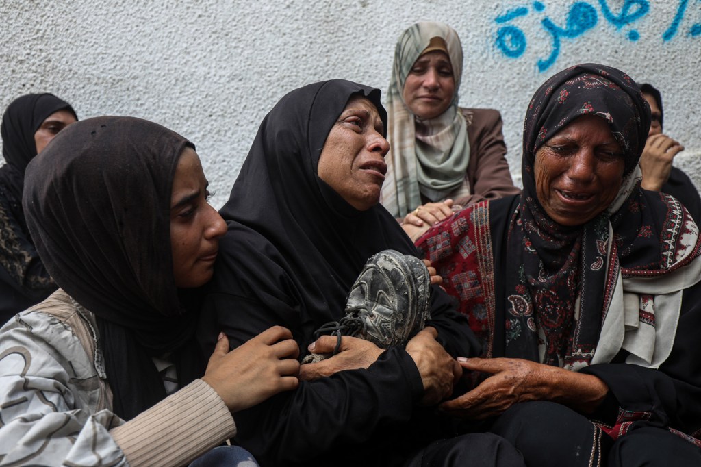 The mother of Khaled Al-Shinbari, a Palestinian teenager killed by Israeli fire while seeking aid in northern Gaza, holds his shoes during his funeral at Al-Shifa Hospital in Gaza City, on August 28, 2025. Relatives react in grief beside her. Image Credit: Omar Ashtawy/apaimages/IPPG