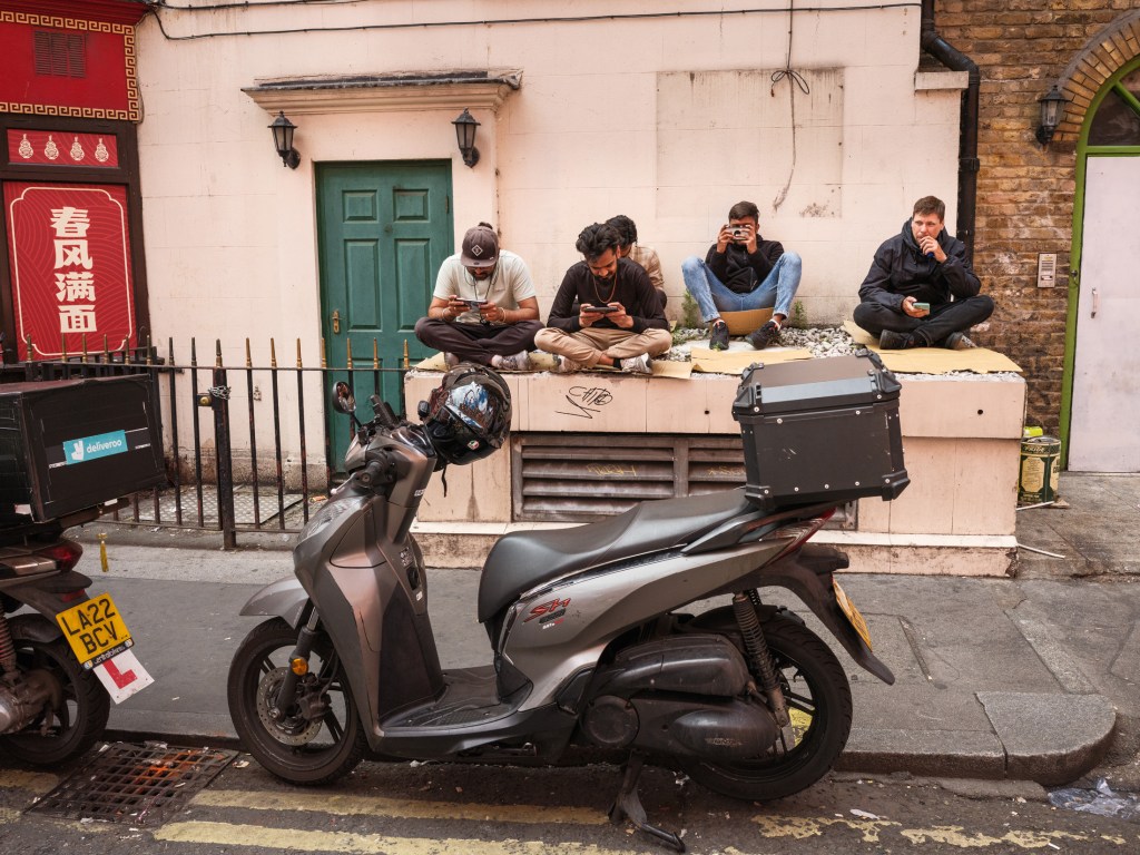 Over 60% of UK adults use a food delivery services, with 80% being accounted for by mobile apps. The forerunners of these services are Deliveroo, Uber Eats, and Just Eat. In the UK, the meal delivery segment is projected to generate £48.21 billion in revenue by the year’s end (2024). Scooter delivery drivers taking a break between jobs. Chinatown, London, UK. July 09, 2025. Photo: Edmond Terakopian