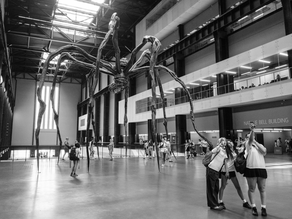 Friends do a selfie by Louise Bourgeois’s giant steel spider sculpture, Maman (1999), as it stands 9-meters high in the Turbine Hall. Tate Modern, London, UK. Processed in LightRoom Classic with the Acros raw profile. This 10,280 ISO has only light colour noise reduction. July 11, 2025. Photo: Edmond Terakopian