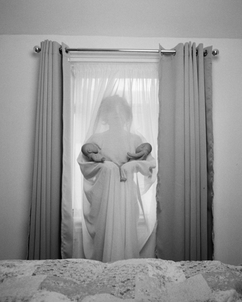 Black and white photograph of a woman behind a net curtain, holding two baby dolls in each arm.