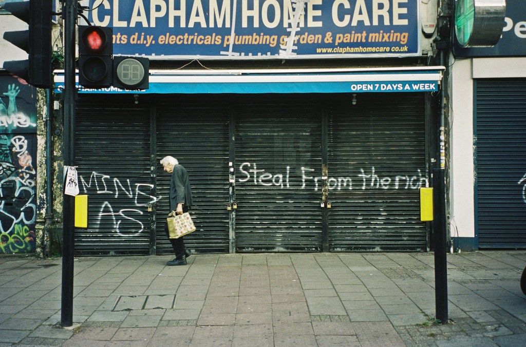 Colour photograph of an elderly person walking along the street past a shuttered shop front that has “STEAL FROM THE RICH” spray-painted in front.