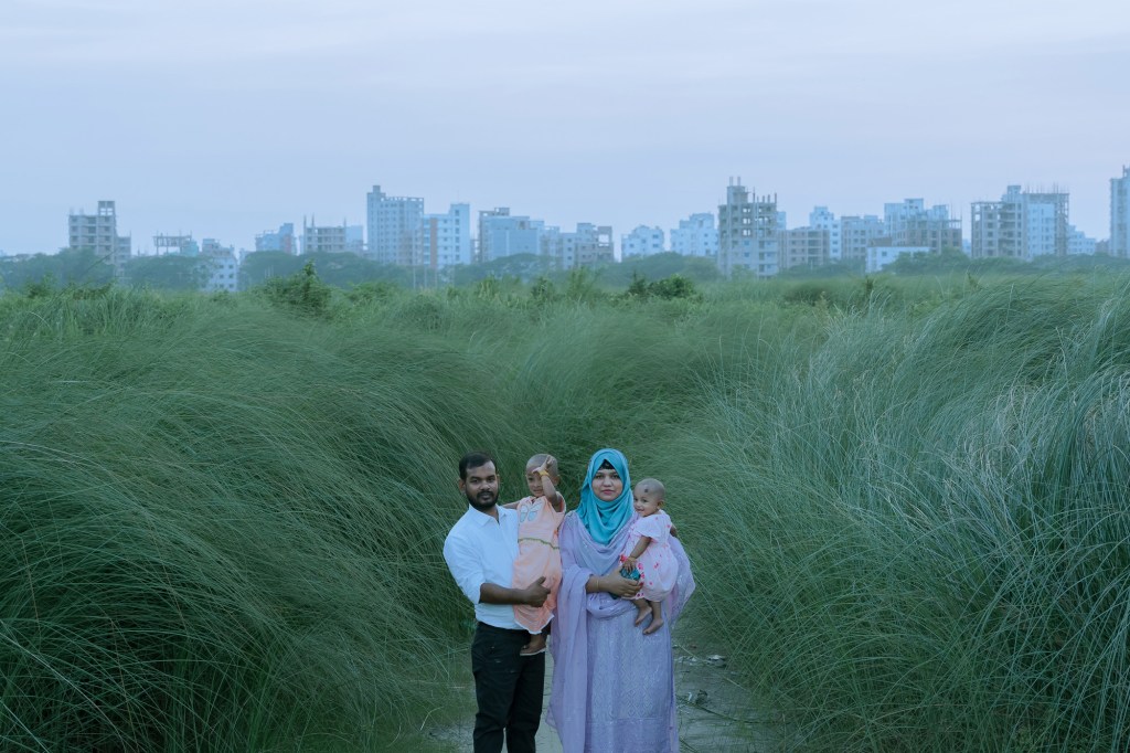 A young family stands in tall grass, children held close, framed by rising buildings. The image evokes resilience and tenderness amid vanishing nature and advancing concrete. The photographer notes, ‘the parents embody adaptation and migration, while their children’s innocence contrasts with looming towers, hinting at hope and uncertain futures.’
