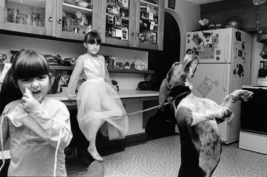 black and white photo of two young girls in the kitchen with a dog that is standing on its back legs