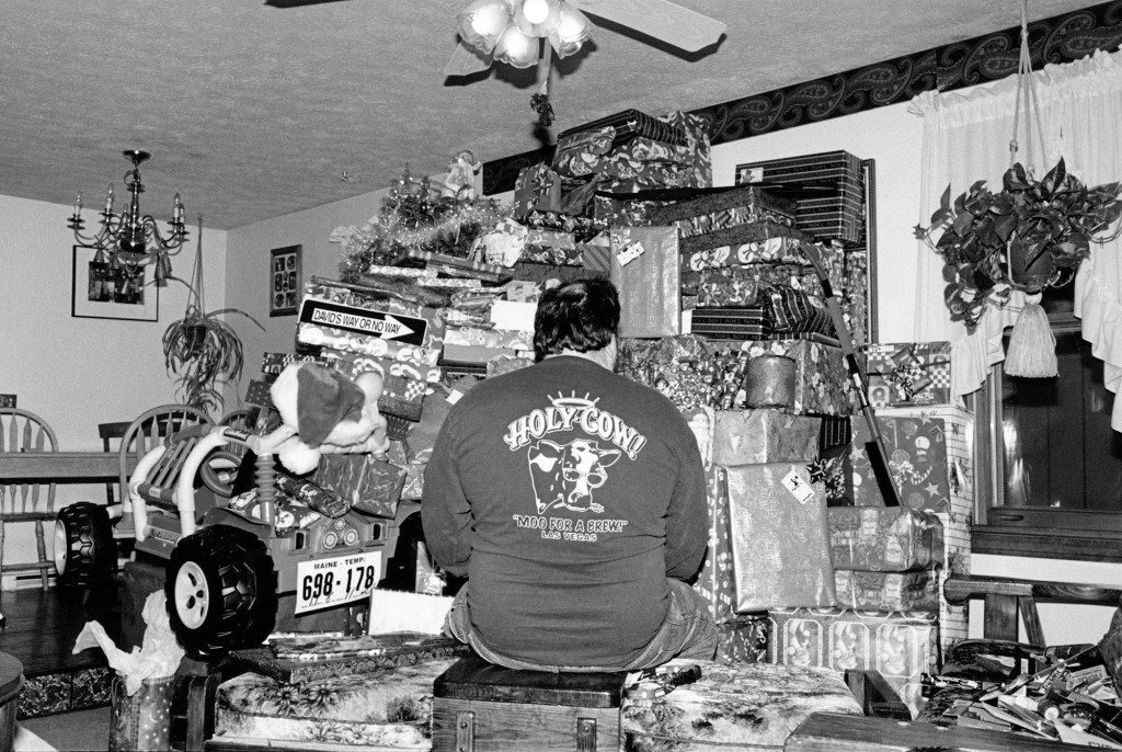 black and white photo of the back of a man wearing a shirt that says Holy Cow sitting in front of a stack of Christmas presents