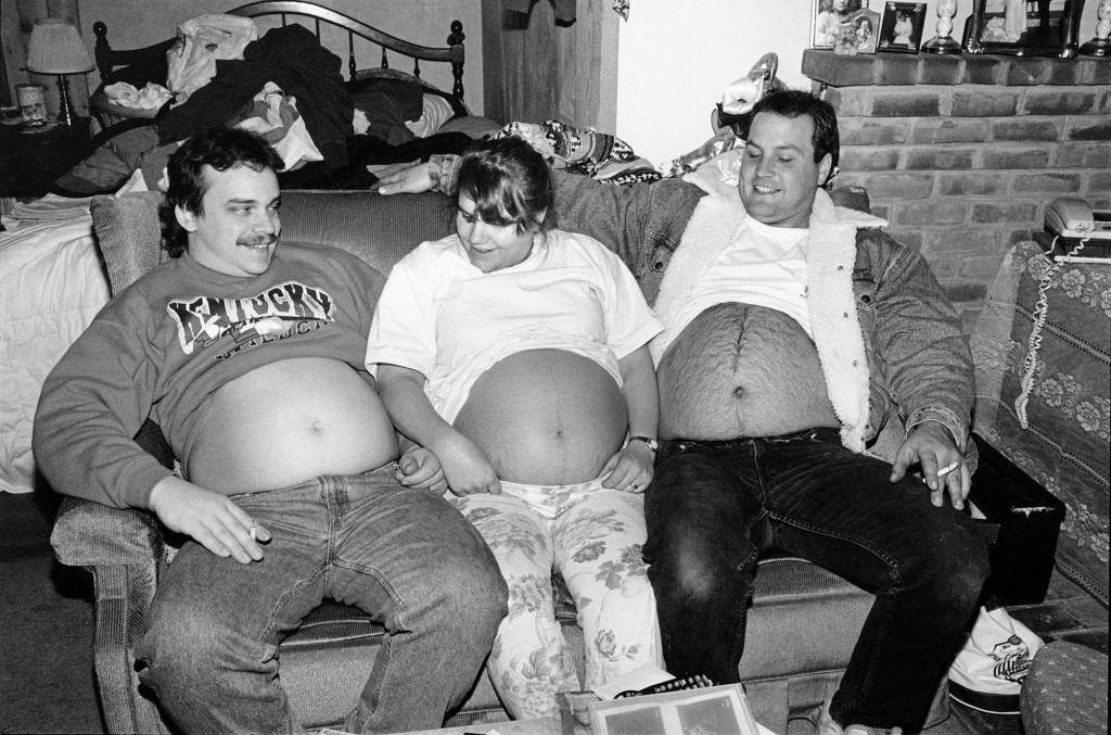 black and white photo of a pregnant woman showing her belly with two men sitting on either side of her also showing their bellies