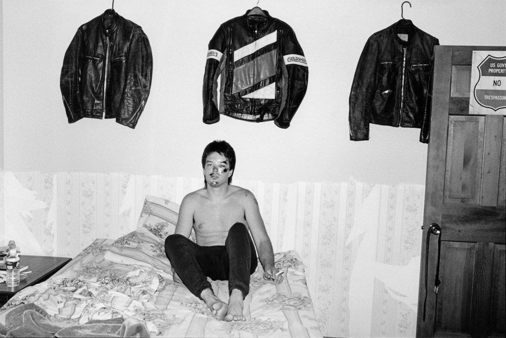 black and white photo of a man sitting on a bed with 3 biker jackets hanging on the wall behind him