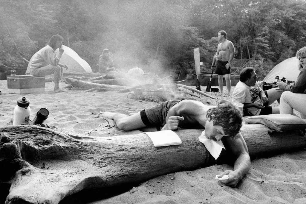 Black and white photo of a group of people at the beach with one passed out over a tree trunk