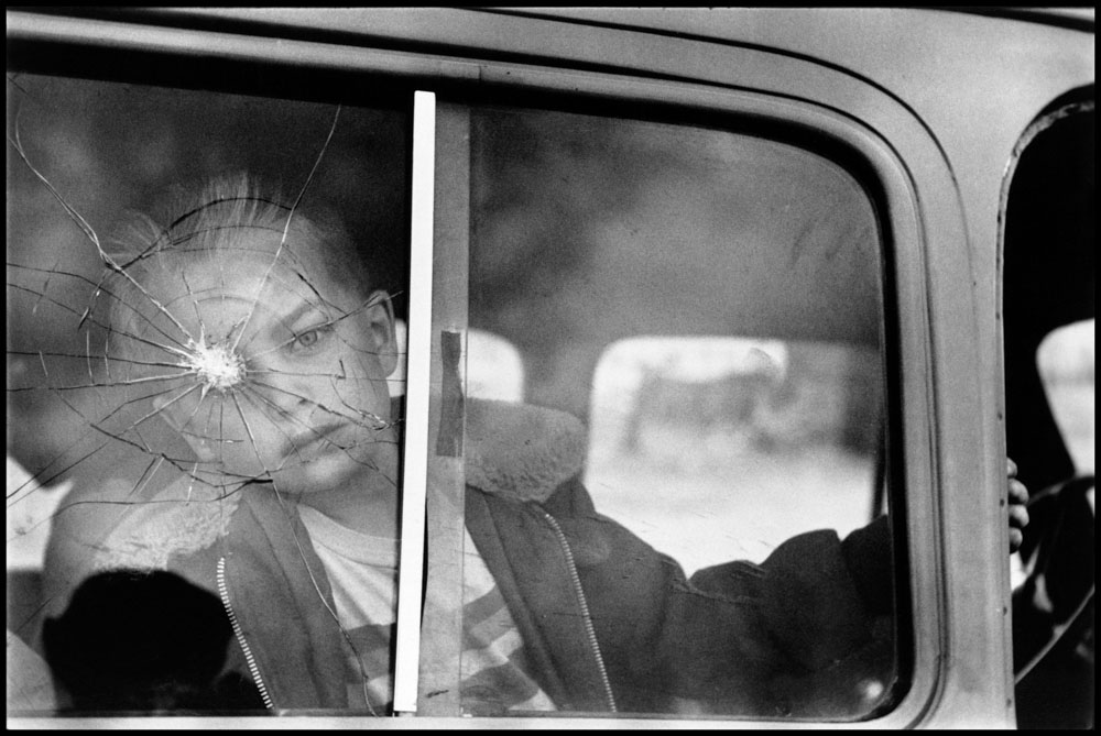 A black and white image of a child looking through a car window which has been shattered by a gunshot