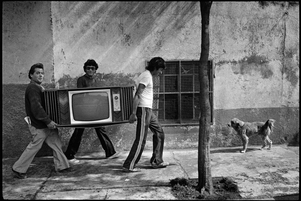 A black and white image showing people carrying a TV through the streets of Mexico