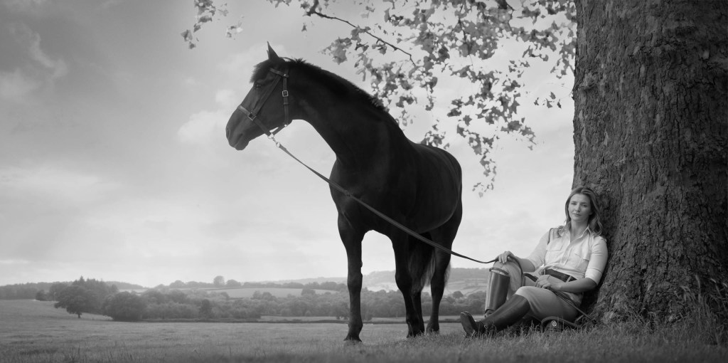 black and white photo of a woman seated on the ground while a horse stands next to her