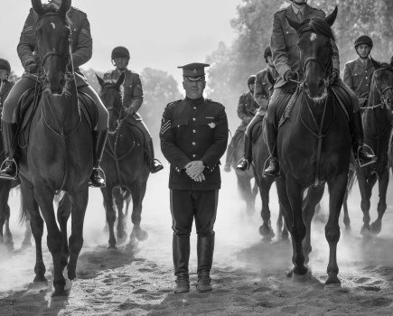 black and white photo of a man standing while mounted horses pass around him