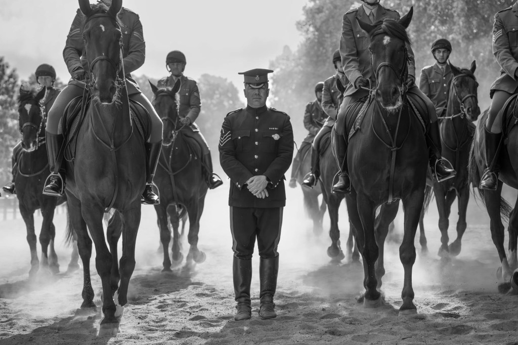 black and white photo of a man standing while mounted horses pass around him