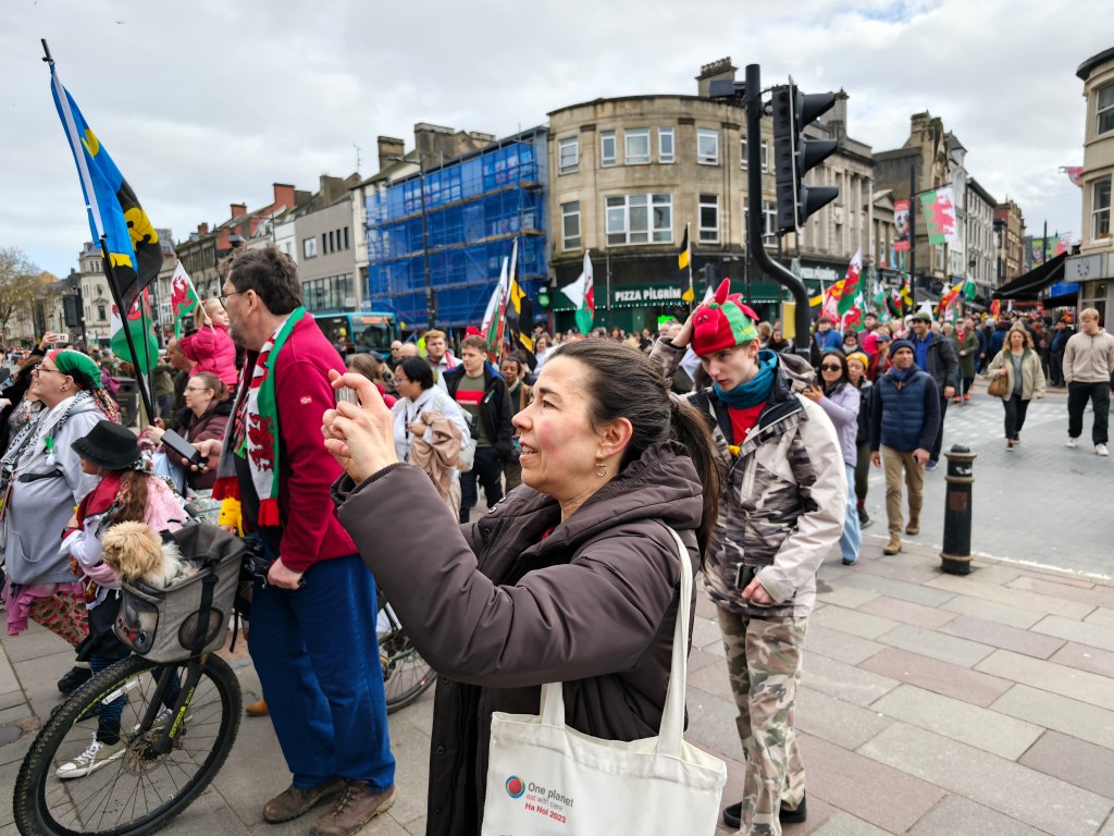 A street scene showing lots of people gathered around some buildings as part of a parade