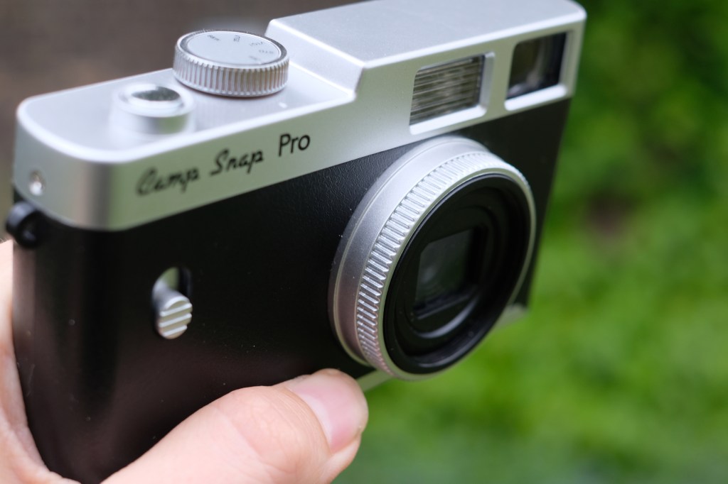 Photograph of the Camp Snap CS-PRO digital compact camera held at an angle facing down, with textured lens ring visible