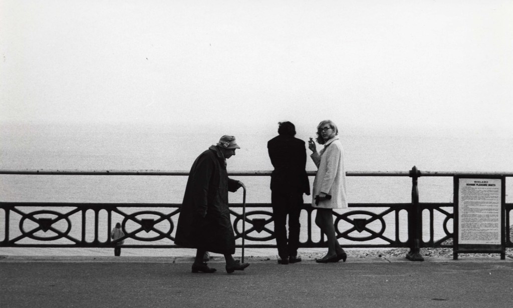 Black and white street photography by Dorothy Bohm of a young couple and an elderly woman with a cane on a seaside pier, featured in the About Women exhibition at Farleys House.