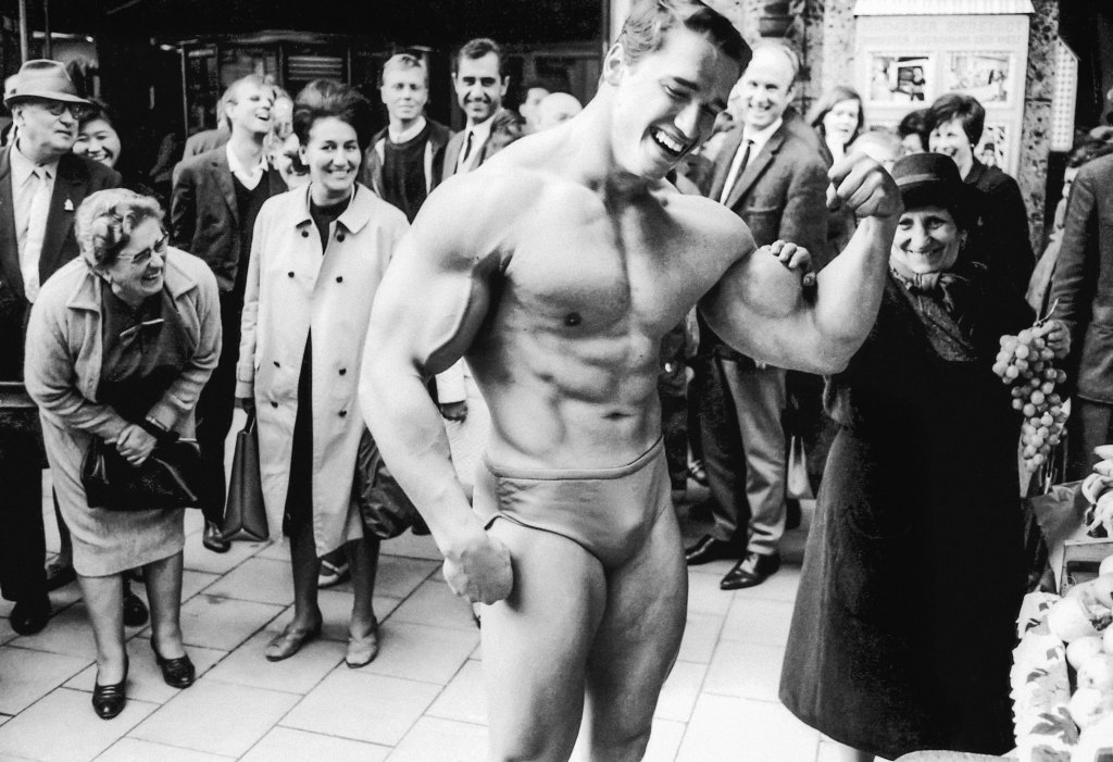 black and white image of Arnold Schwarzenegger in underwear on busy street, with a lady admiring his muscles