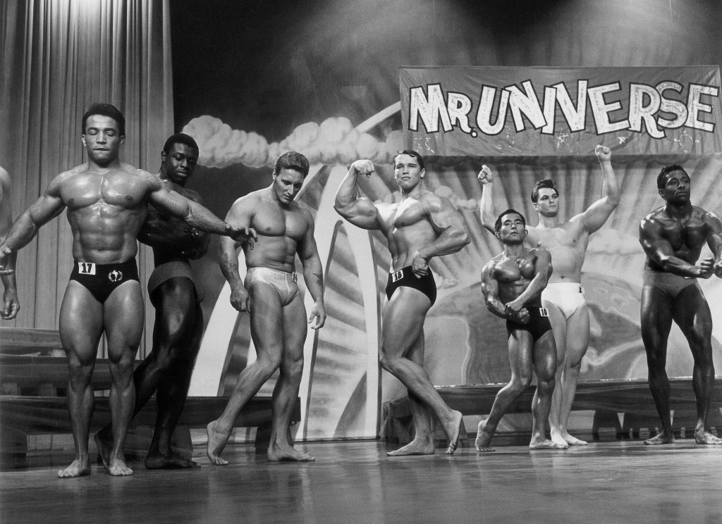 black and white image of Arnold Schwarzenegger and other bodybuilders posing on Mr Universe stage